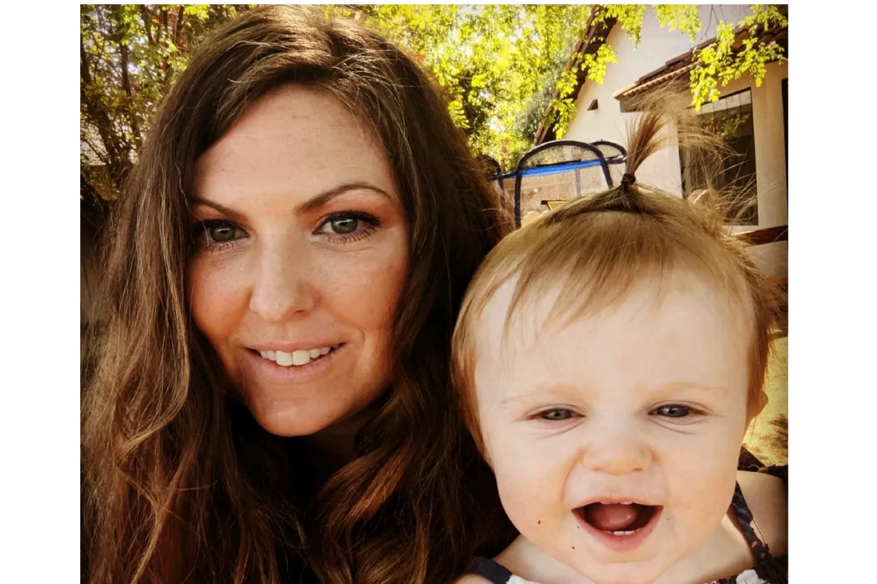 A woman and a baby smiling outdoors under sunny trees.
